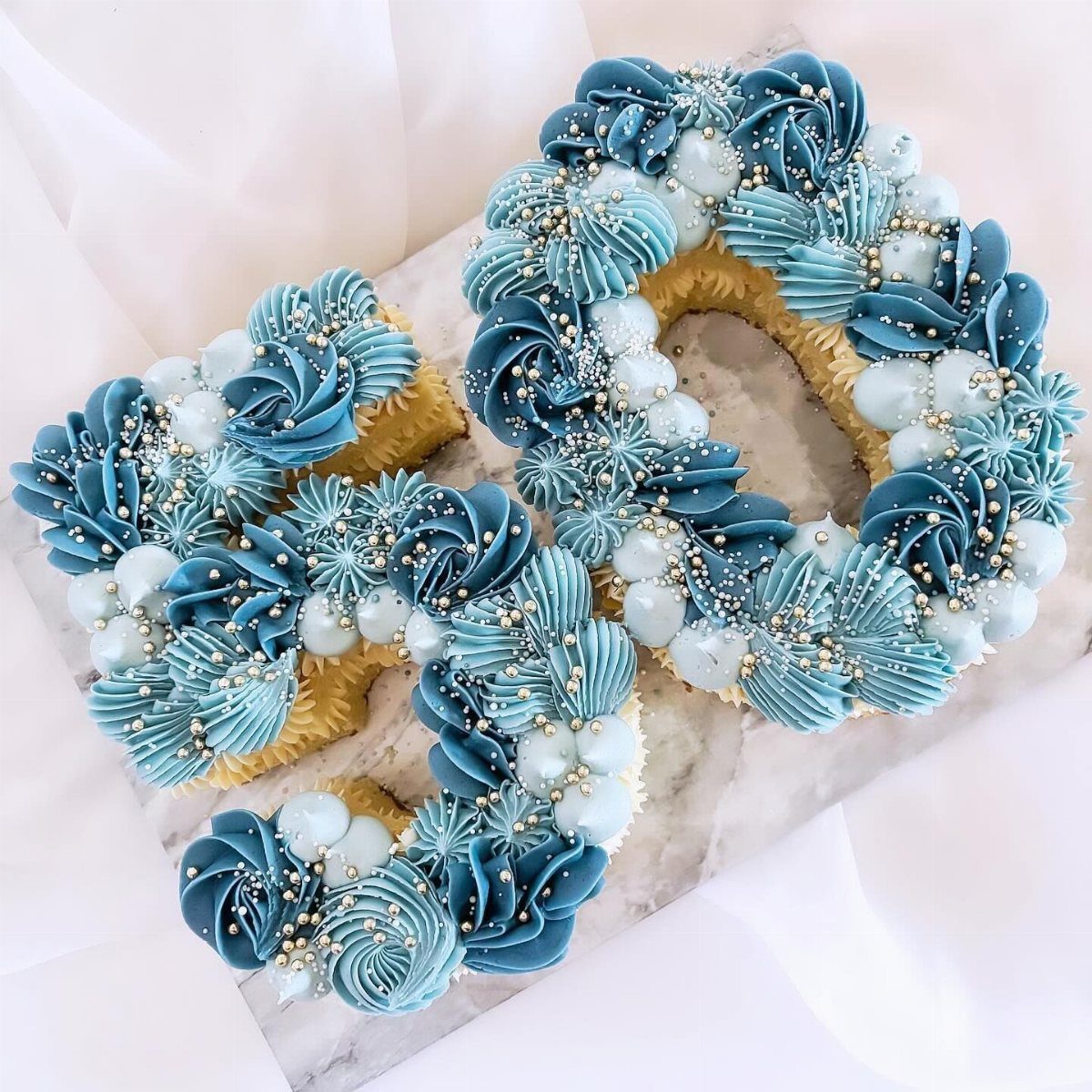 Number 50 cake decorated with blue frosting swirls, white pearls, and gold sprinkles.