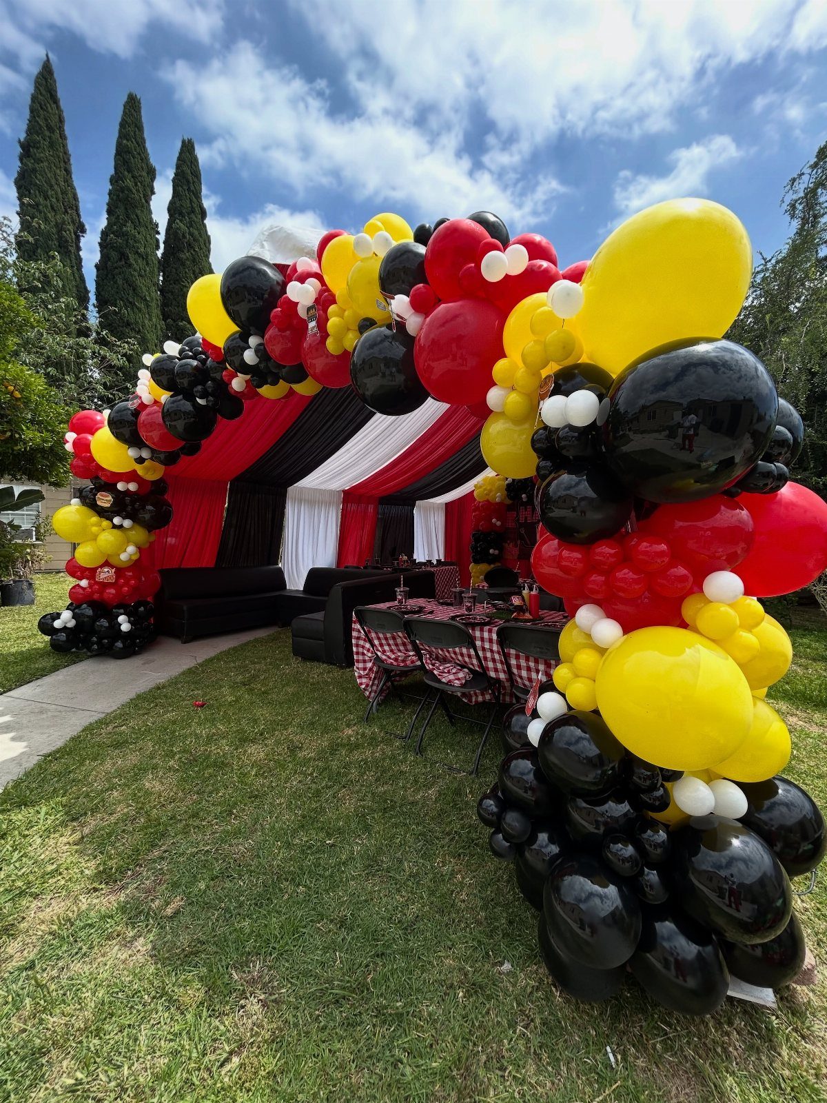 Red, yellow, and black balloon arch framing a party table outdoors.