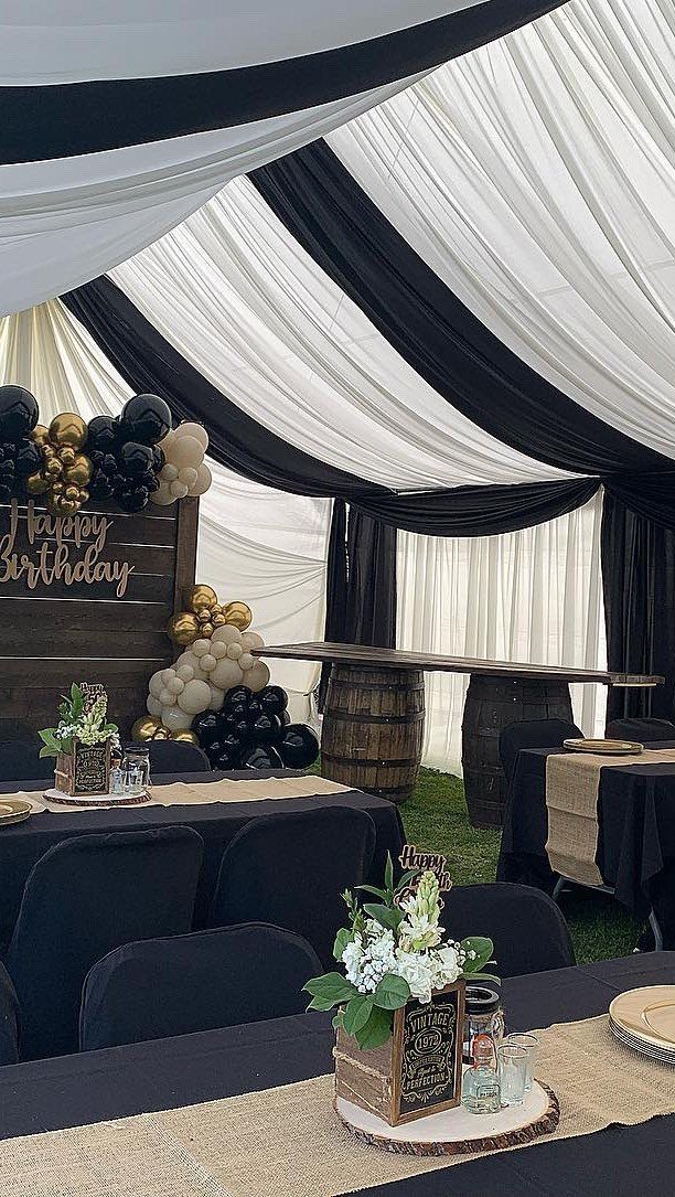 Birthday party decorations with black and white draping, gold balloons, and rustic barrel tables.