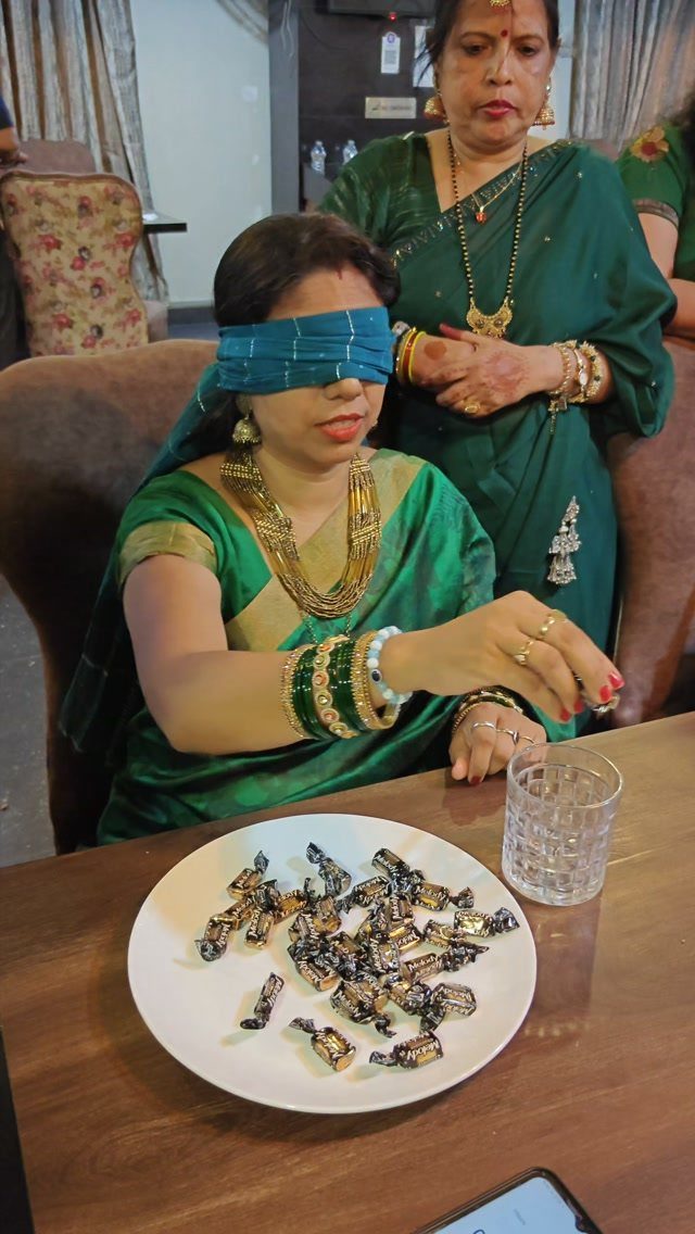 Blindfolded woman in green saree reaching for candies on a white plate