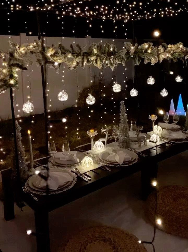 Outdoor dinner table with white settings under glowing string lights and frosted greenery.