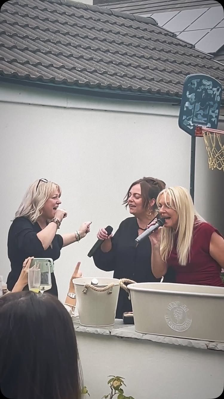 Three women singing into microphones during an outdoor party celebration.