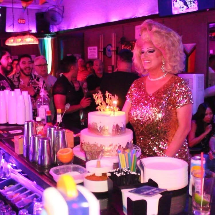 Drag queen in gold sequins standing by a birthday cake at a bar