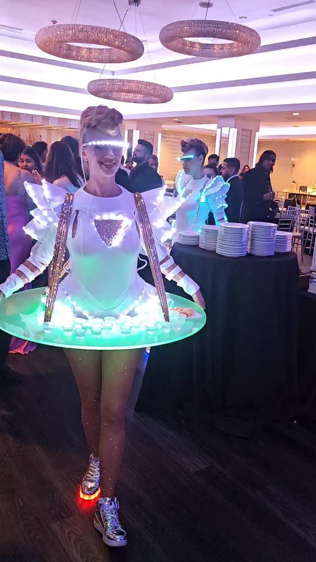 Woman in glowing futuristic costume serving drinks on a waist-mounted tray at a party