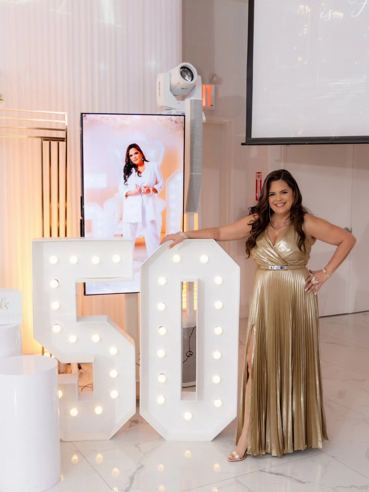 Woman in a metallic gold pleated dress posing by large illuminated 50 numbers.