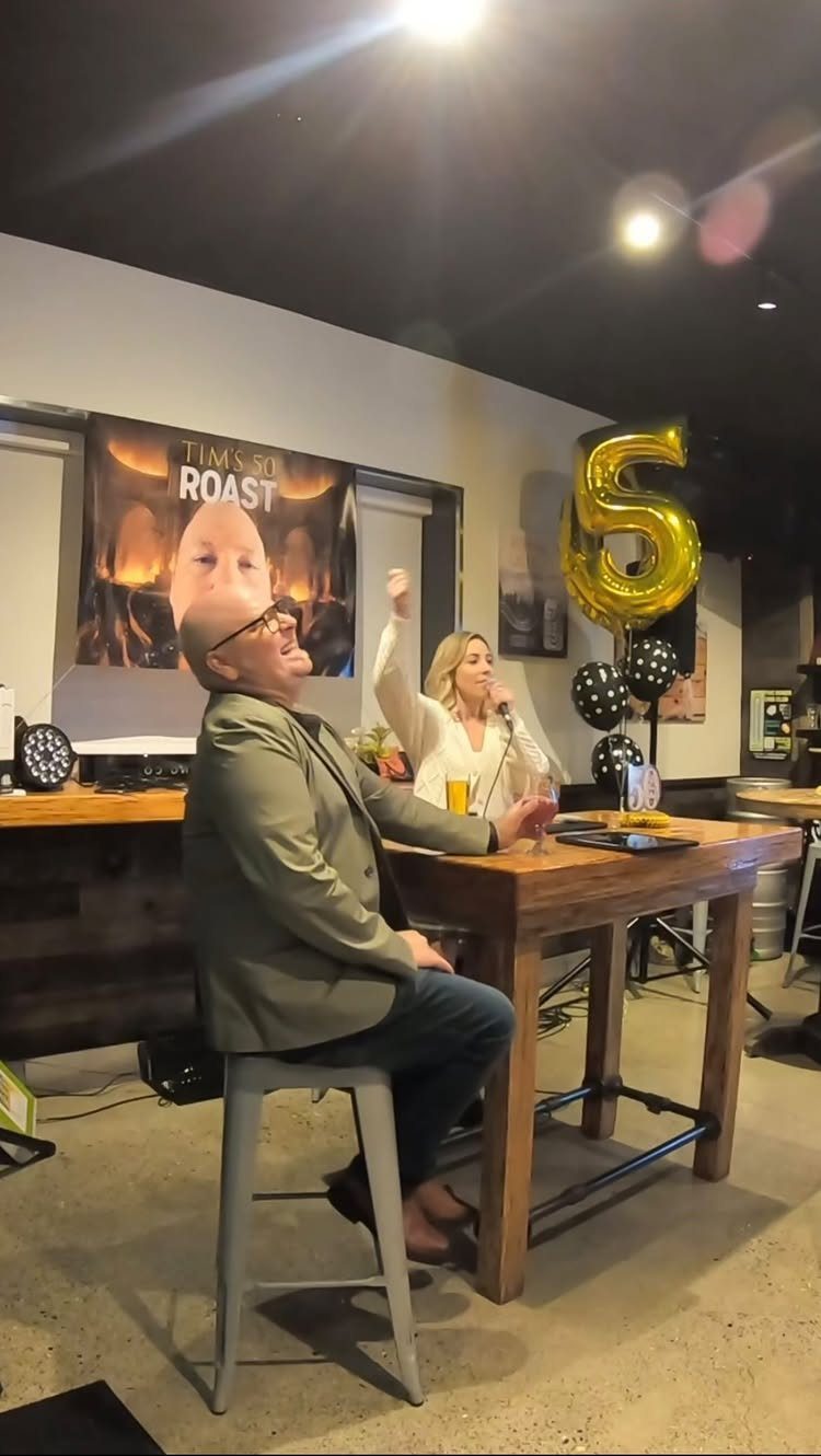 Man laughing while a woman speaks into a microphone during a roast party.