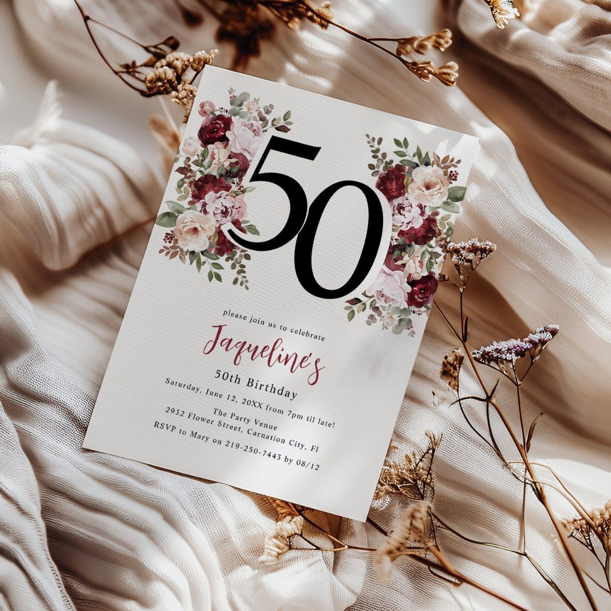 Floral 50th birthday invitation card resting on beige fabric with dried flowers.