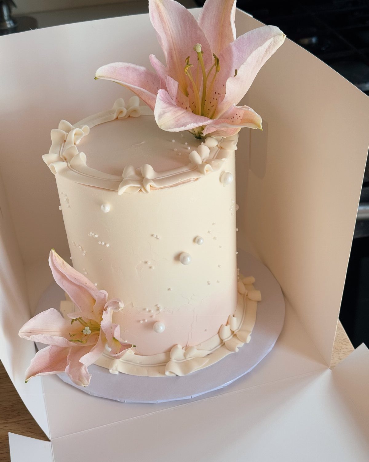 White frosted cake decorated with pink lilies and small white edible pearls.