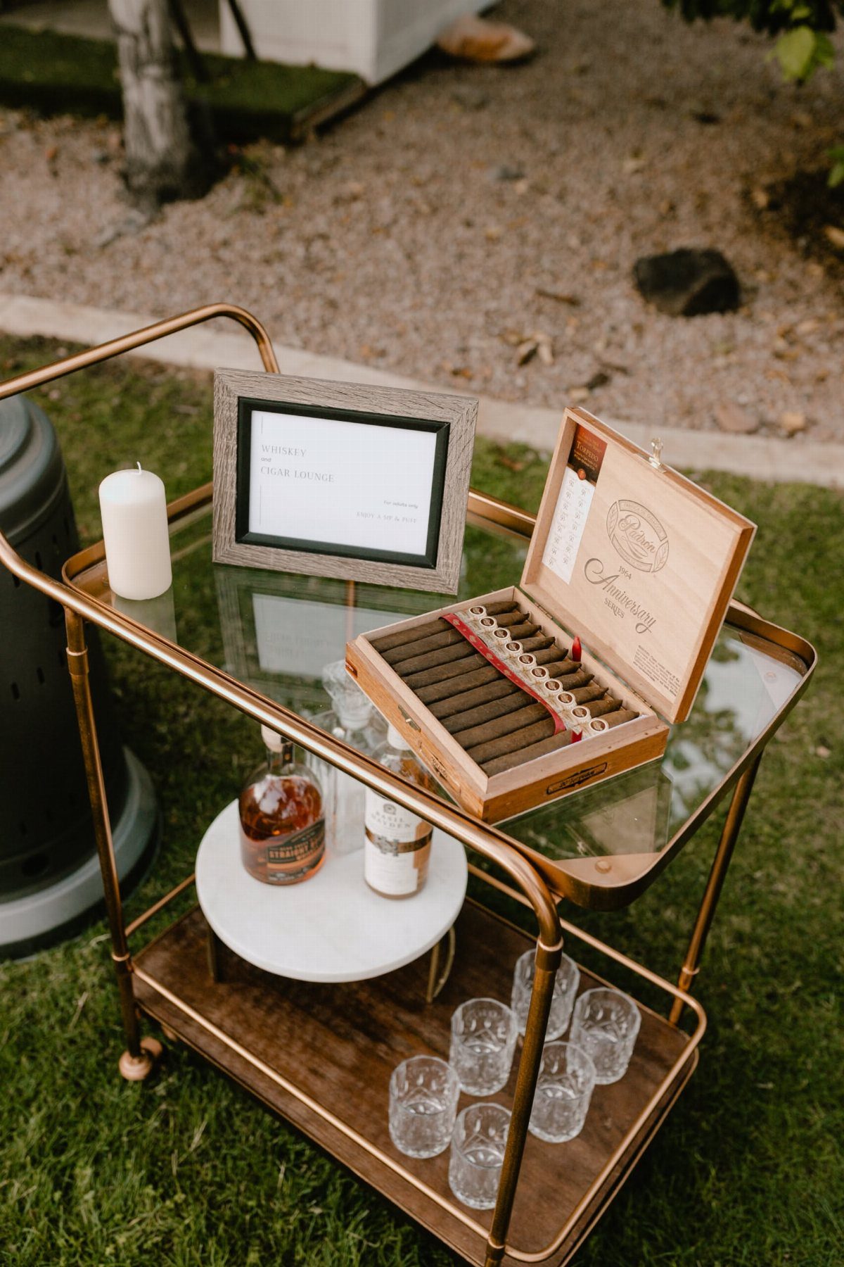 Gold bar cart with a cigar box, liquor bottle, and glasses on grass.