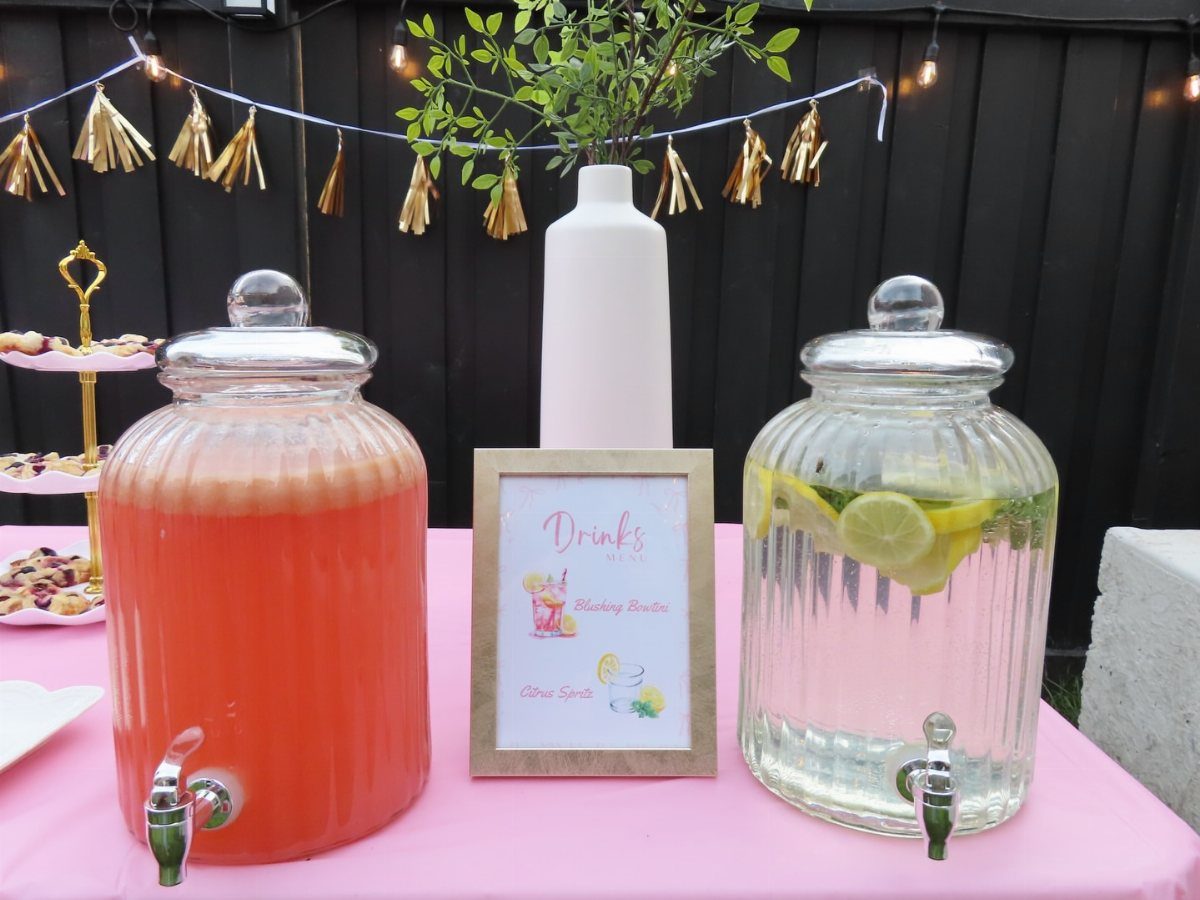 Beverage dispensers with red and lemon water on a pink table outdoors.