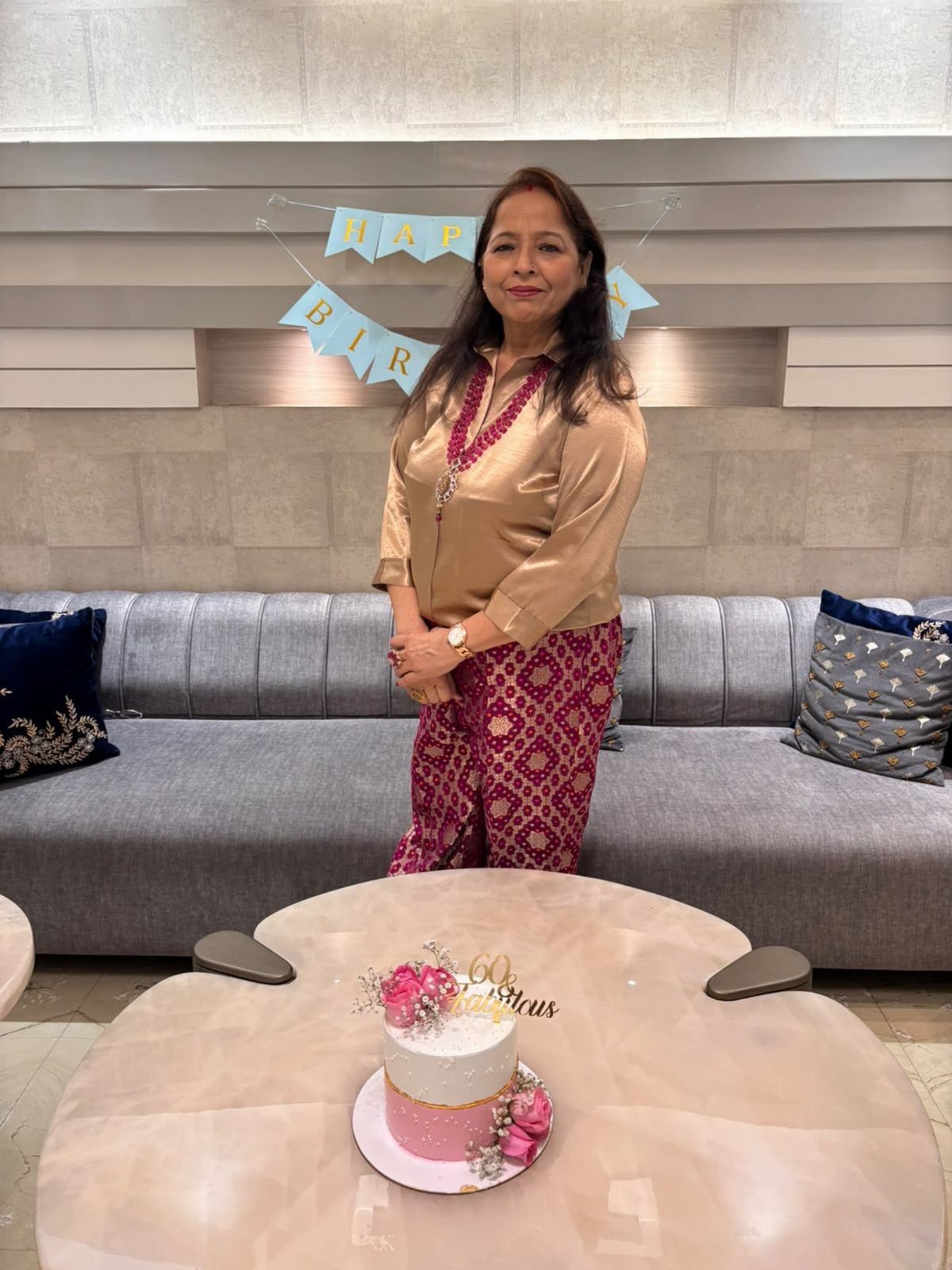 Woman in gold blouse and maroon trousers standing by a birthday cake