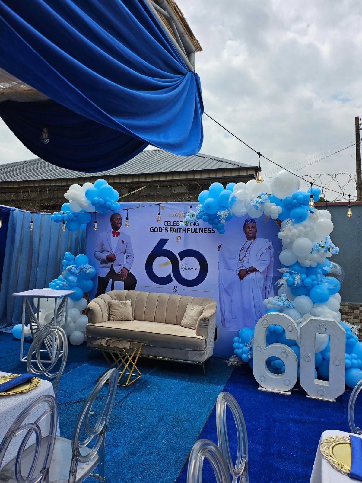Outdoor 60th birthday party setup with blue balloons, beige sofa, and large 60 numbers.