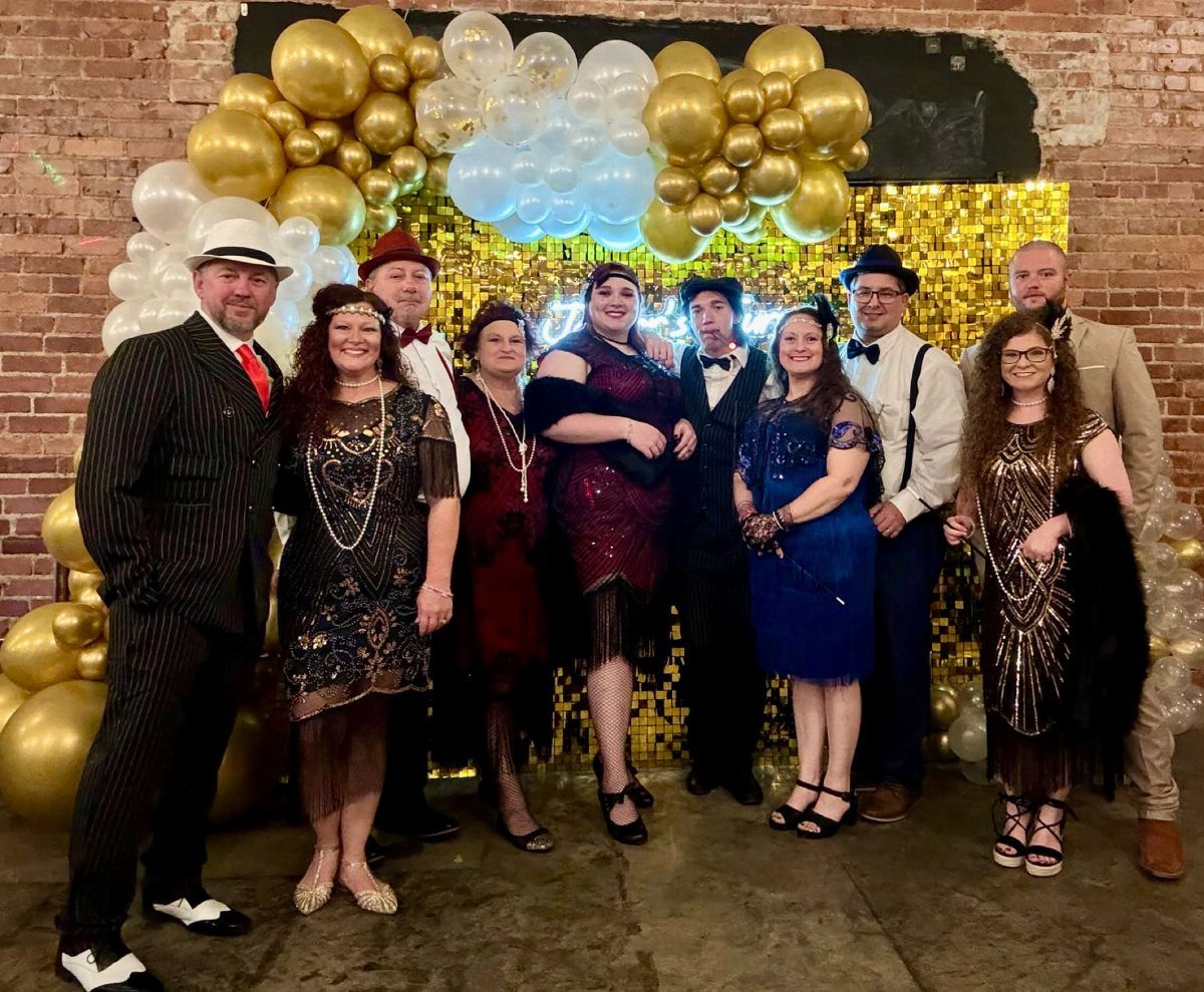 Group of people wearing 1920s flapper dresses and suits before a gold backdrop.