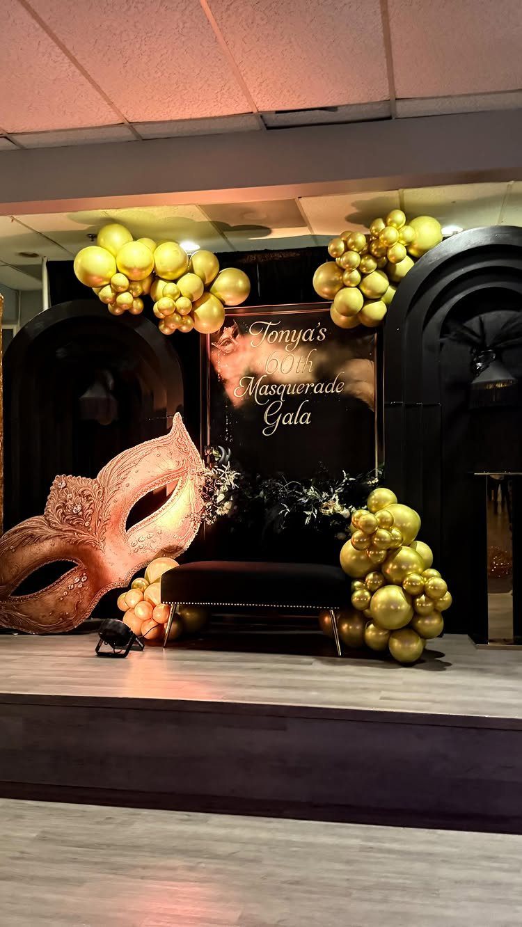 Gold balloons and a large mask decorate a masquerade gala party backdrop.