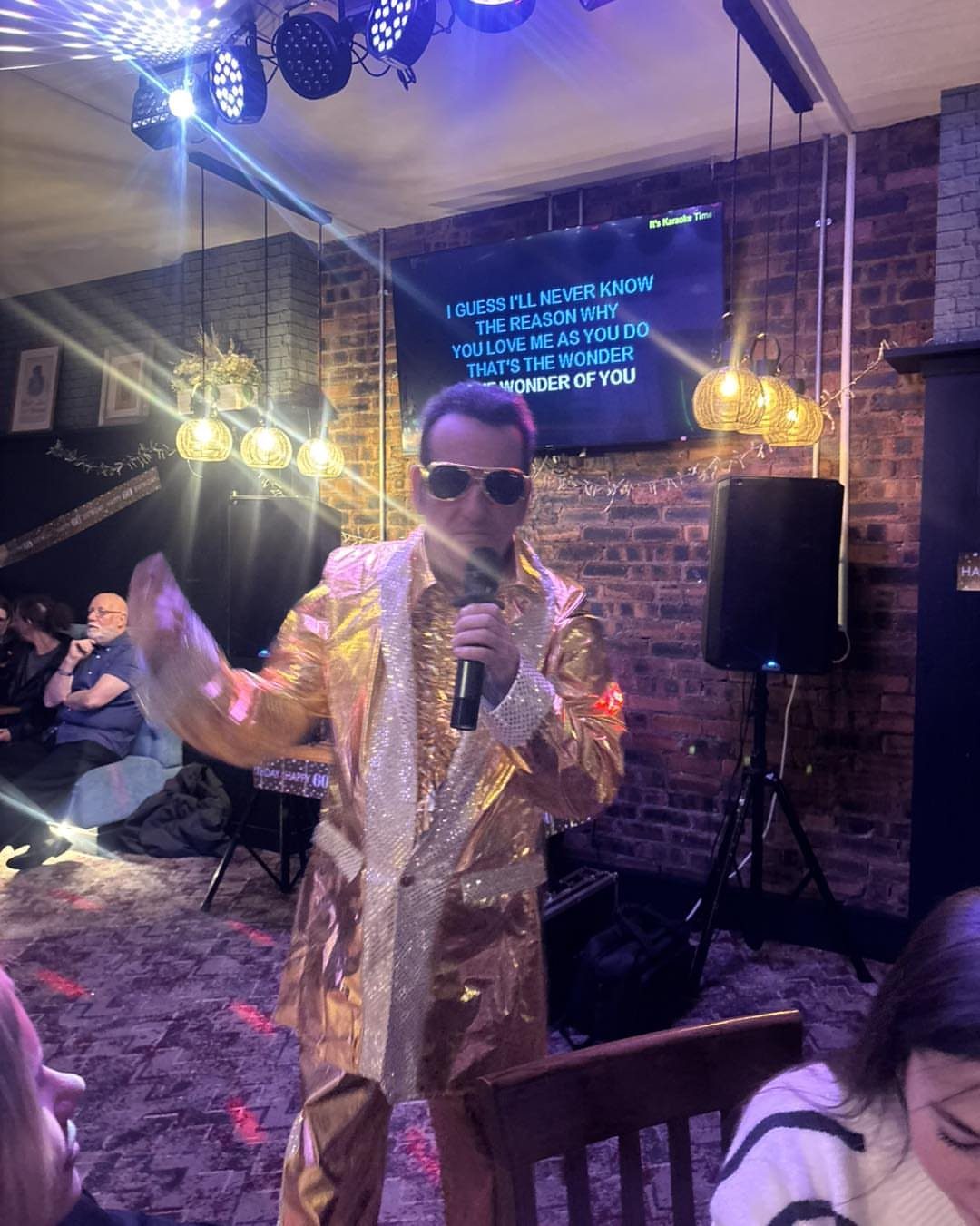 Man in gold sequined suit singing into a microphone with lyrics on screen.