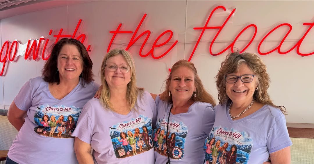 Four women in matching purple 60th birthday shirts smiling in front of neon signage.