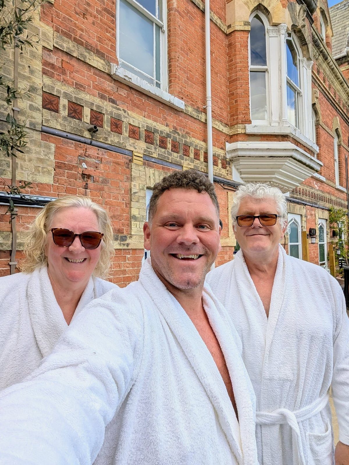 Three adults in white bathrobes and sunglasses smiling for a selfie outdoors.