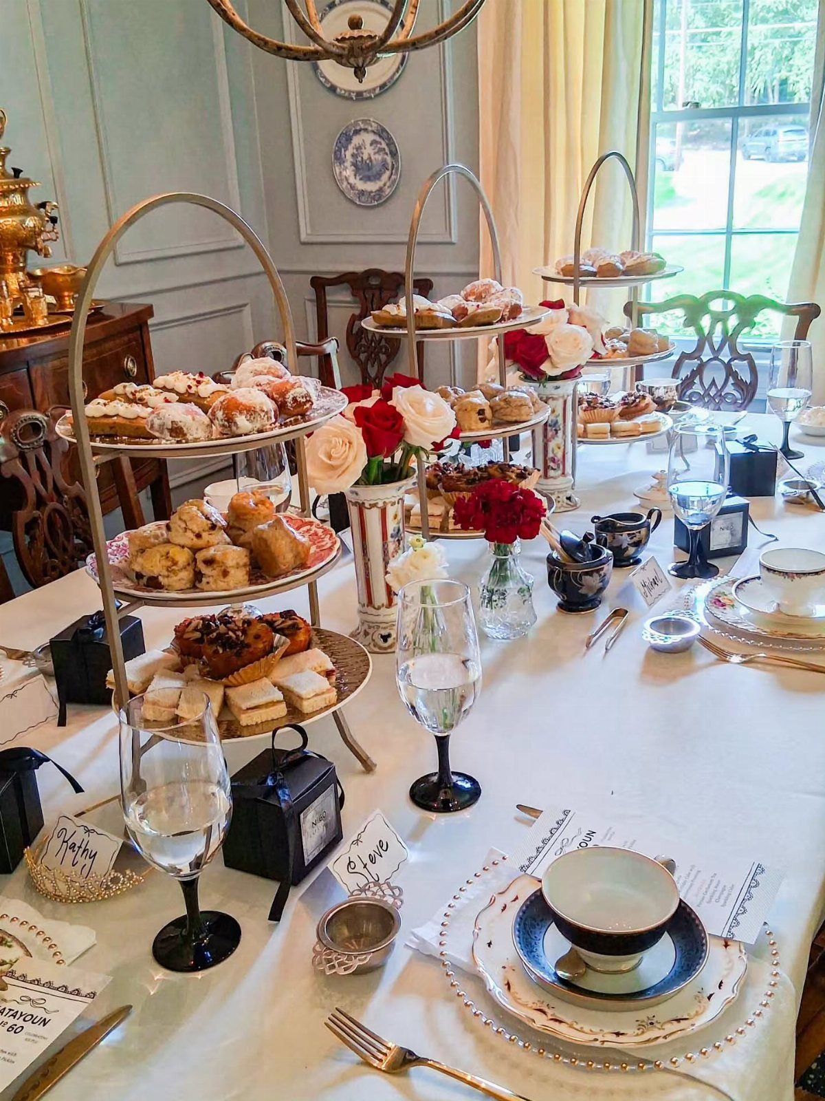 Tiered cake stands with pastries and teacups on a white tablecloth with roses.