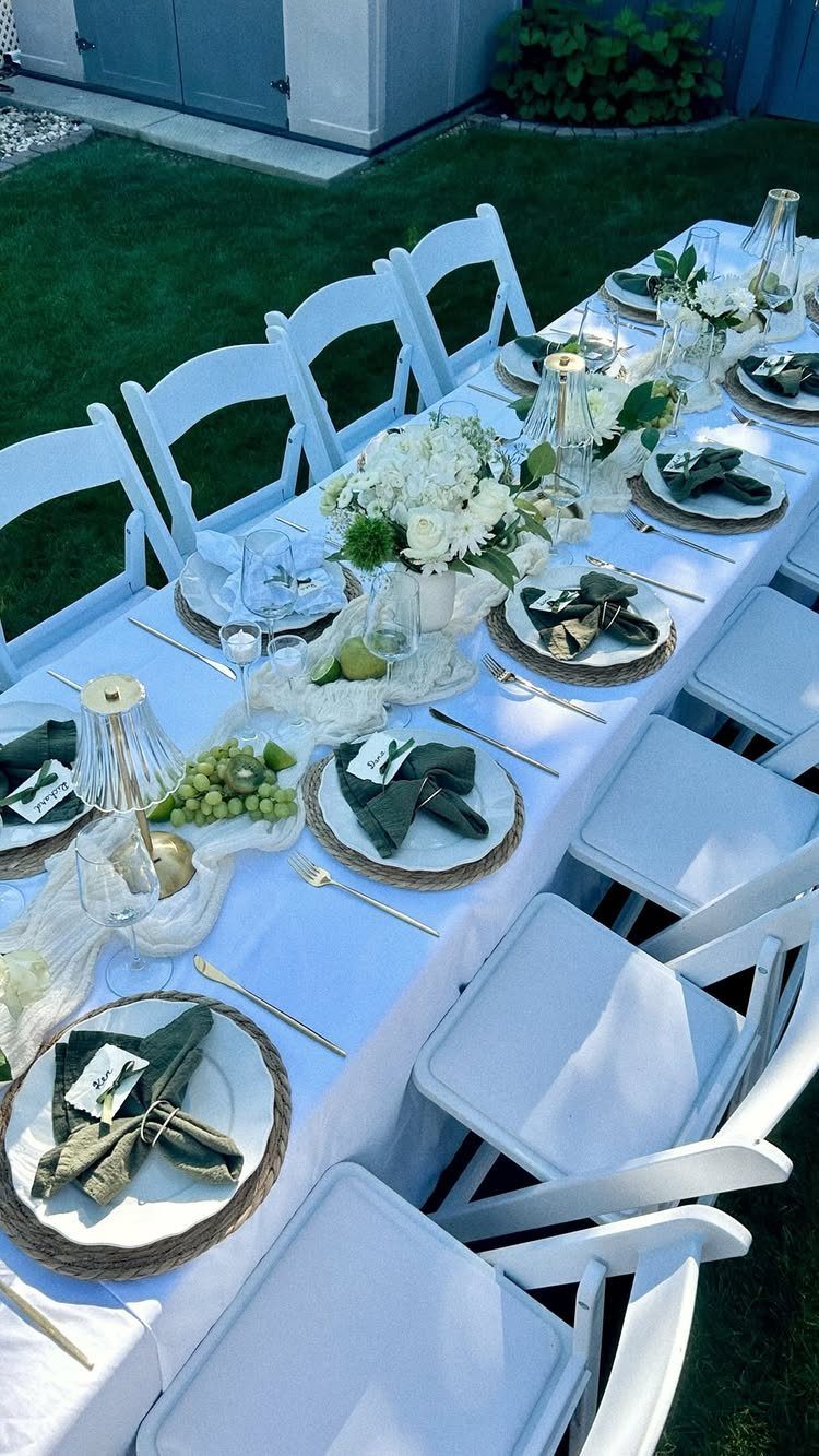 Long outdoor dining table with white linens, green accents, and white floral centerpieces.