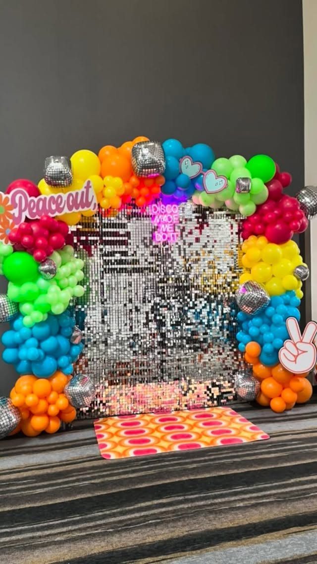 Rainbow balloon arch with disco balls framing a silver sequin party backdrop.
