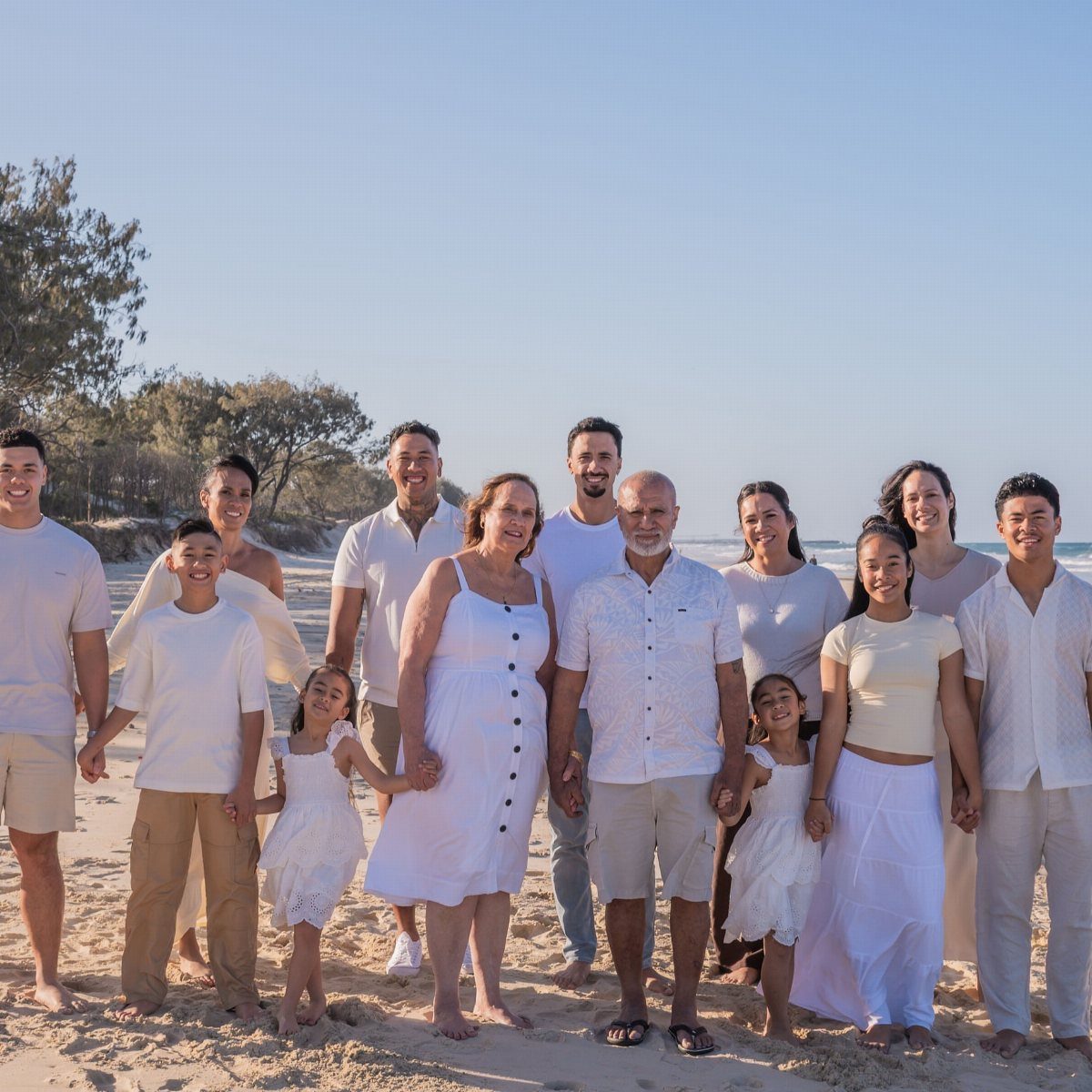 Multi-generational family in white clothing posing together on a sunny sandy beach.