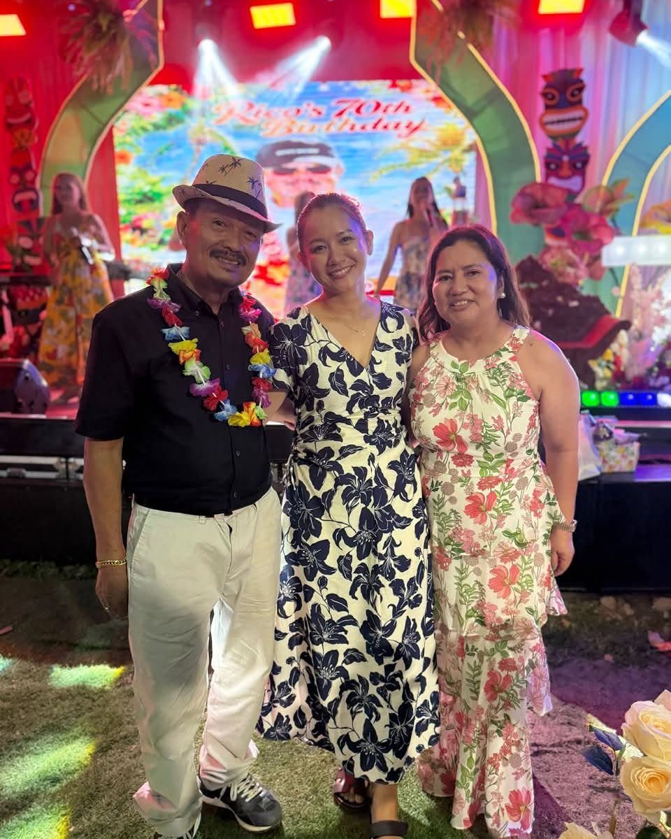 Three people posing at a 70th birthday party with tropical luau decorations.