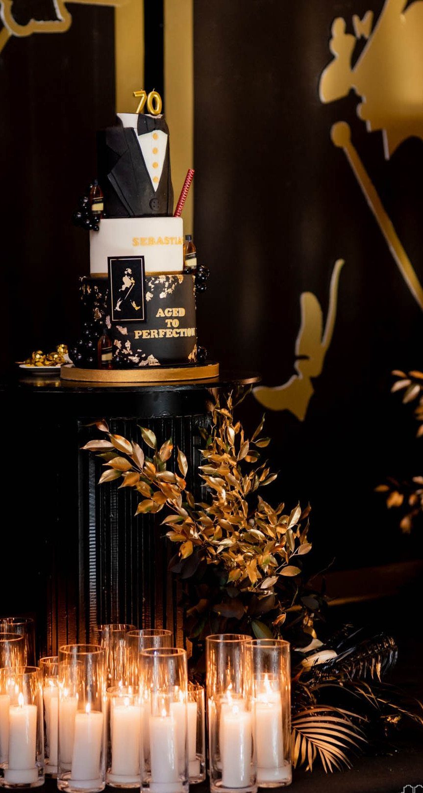 Tuxedo-themed 70th birthday cake with gold topper and glowing white pillar candles.