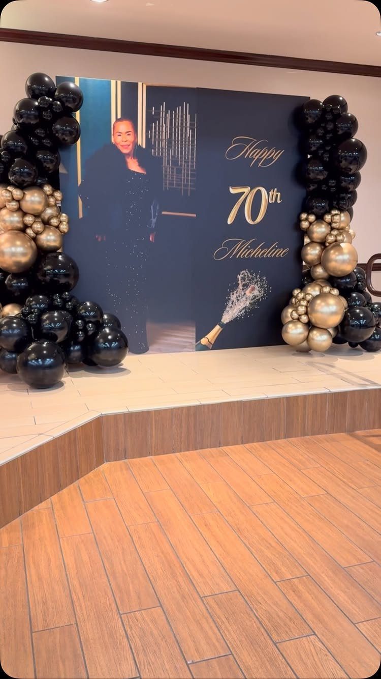 Black and gold 70th birthday backdrop with balloon arches and a woman's portrait.