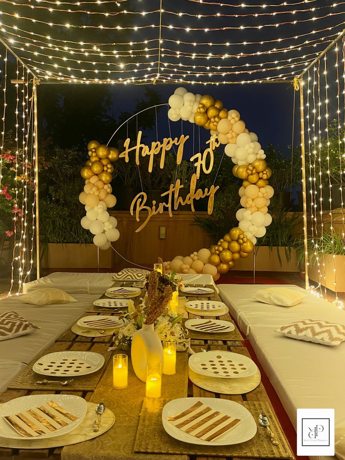Outdoor 70th birthday dinner setup with gold balloons, fairy lights, and floor seating.
