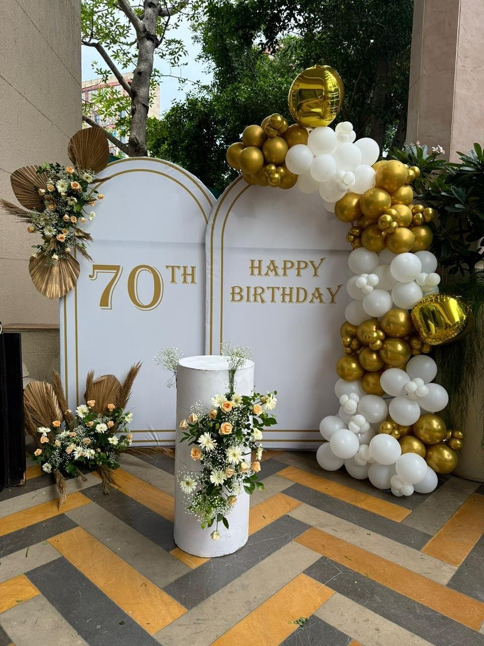 White and gold 70th birthday photo backdrop with balloon arch and floral arrangements.