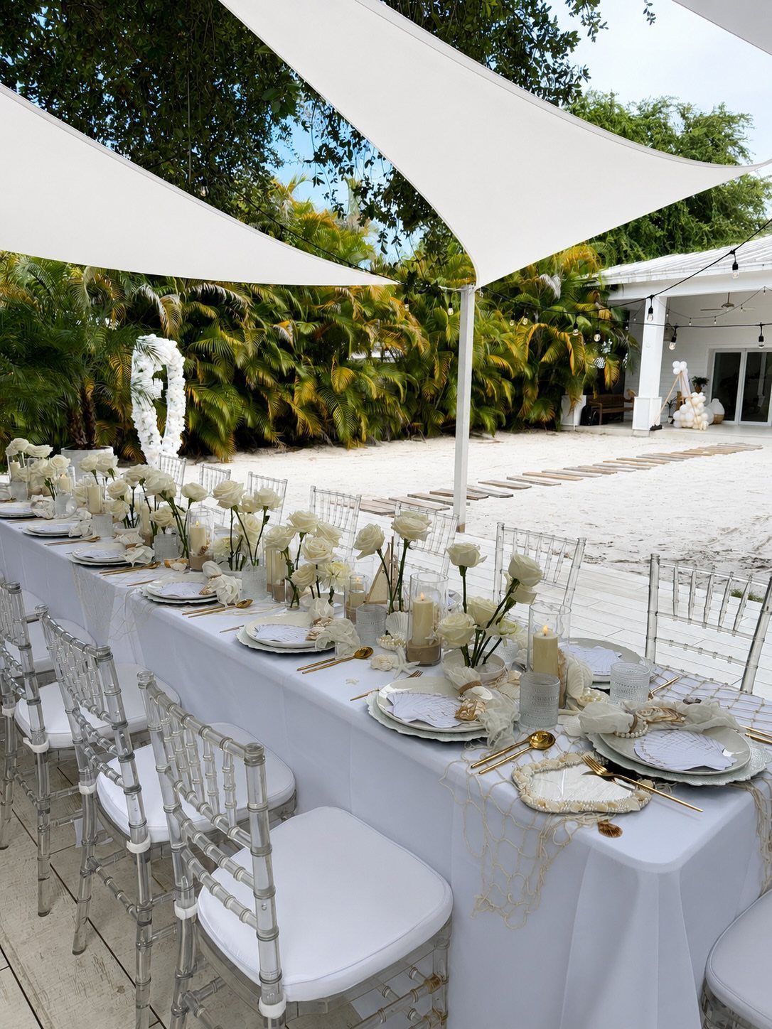 Formal white dining table set on a sandy beach under white sail canopies.