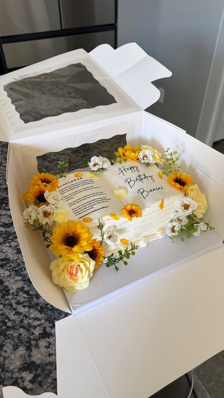 Book-shaped white cake decorated with yellow sunflowers and white blossoms in a box