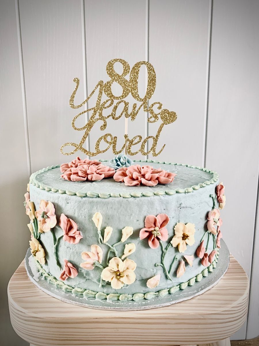 Light blue frosted cake with pink and yellow flowers and gold 80th topper