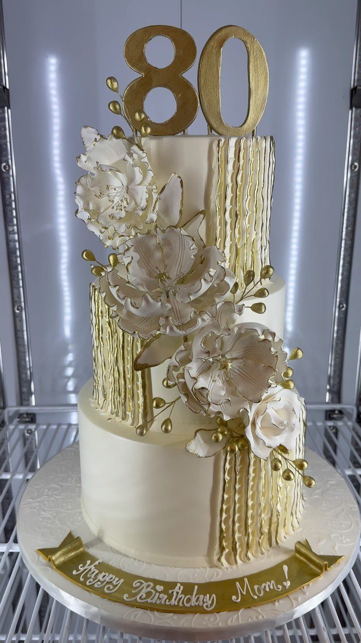 Three-tier white birthday cake with gold drips, white flowers, and gold 80 topper