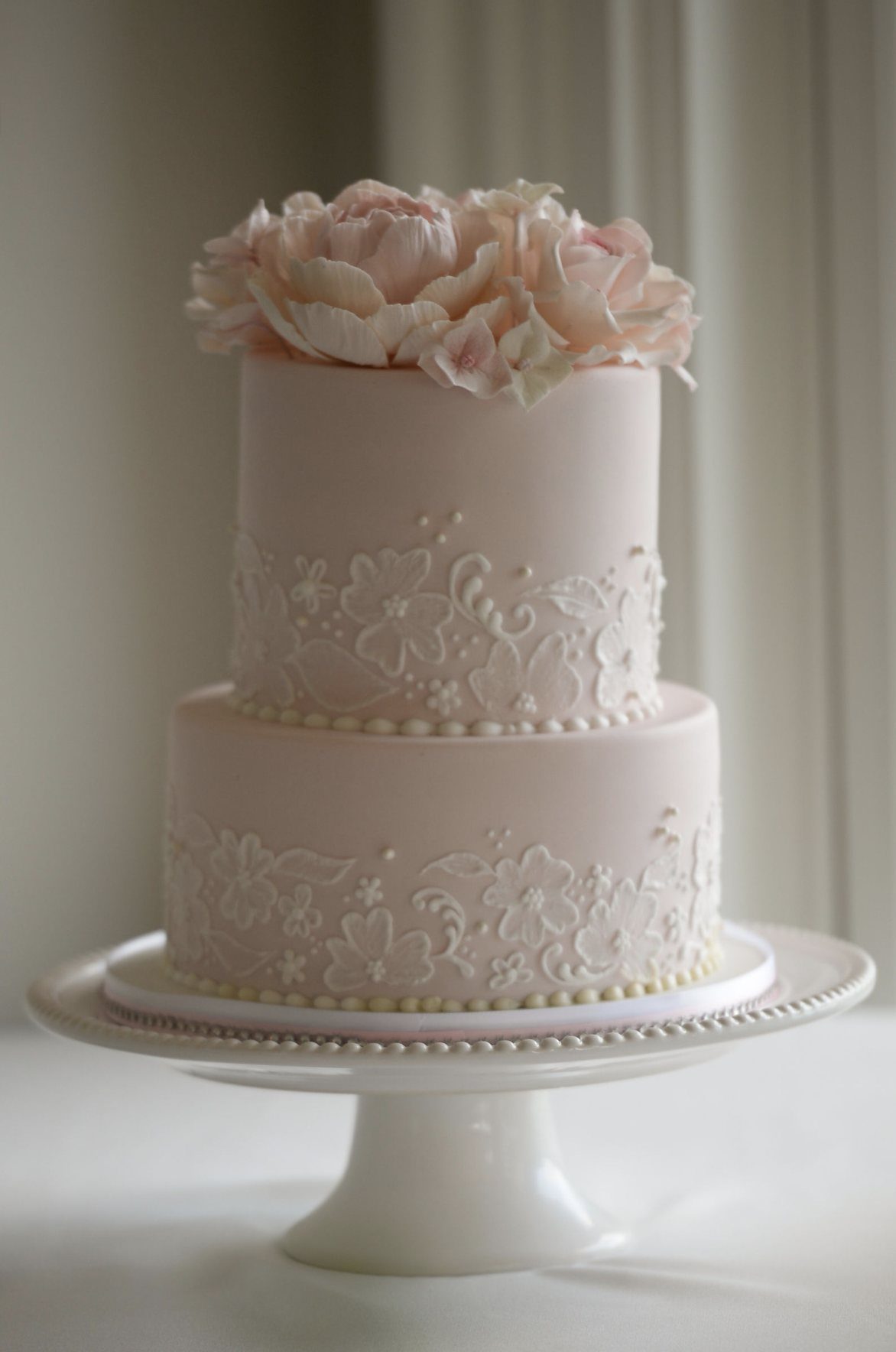 Two-tier pale pink cake with white floral lace piping and pink flower topper.