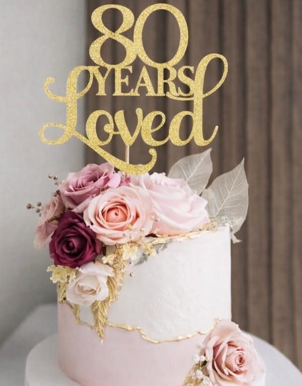White and pink frosted cake with pink and purple roses and gold topper.