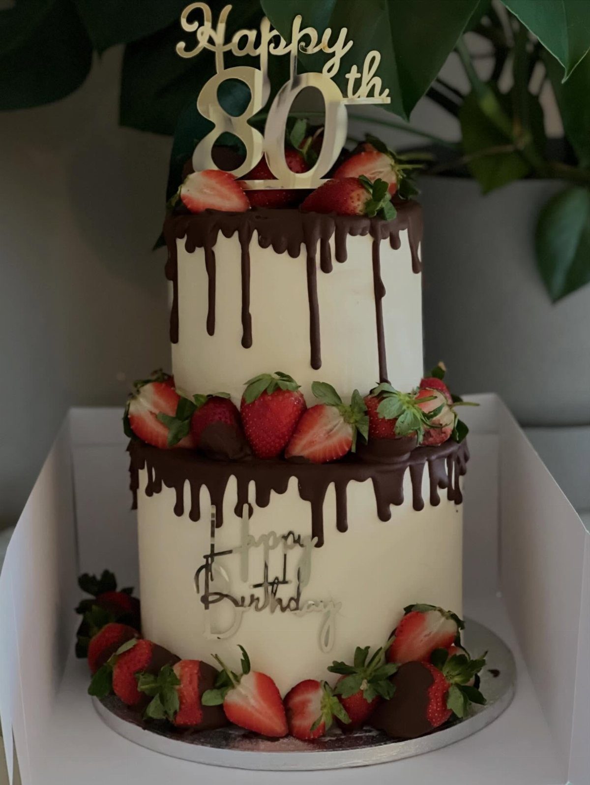 Two-tier white cake with chocolate drips, fresh strawberries, and a gold 80th topper.