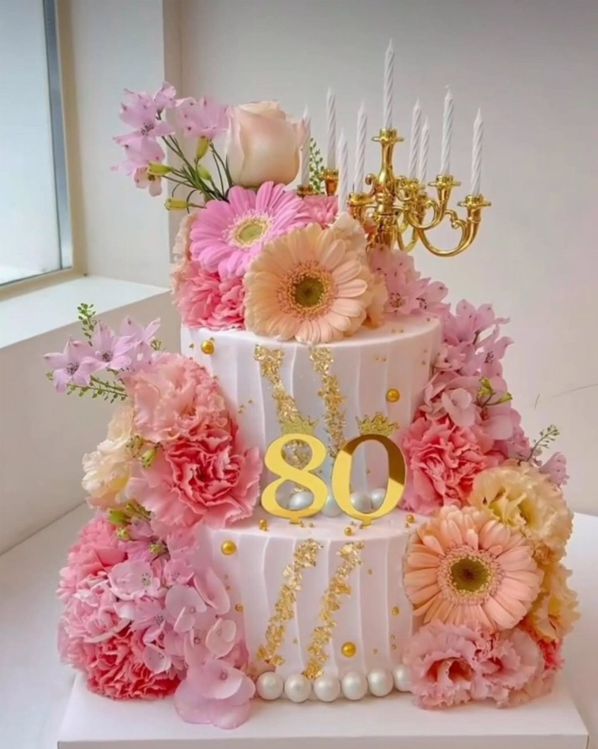 Two-tier white cake with pink flowers, gold 80 topper, and gold candelabra.