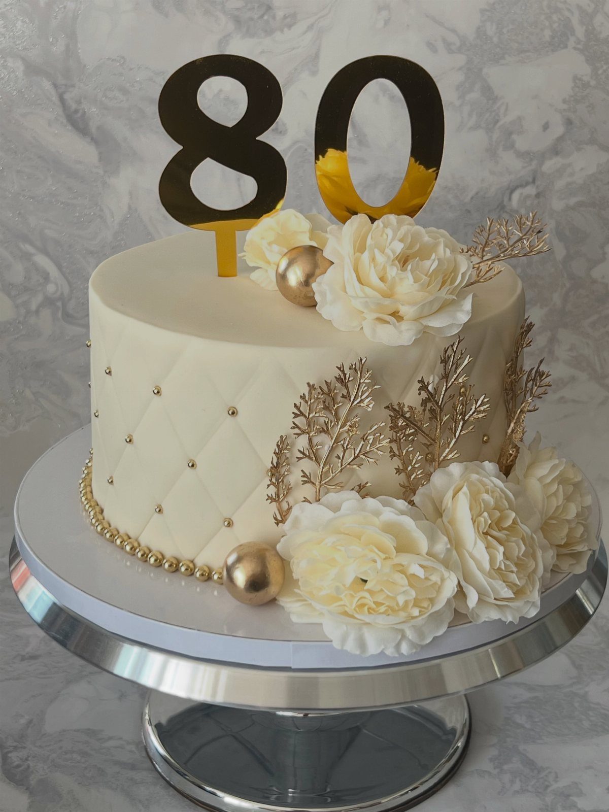 White quilted cake with gold 80 topper, white flowers, and gold leaf accents.