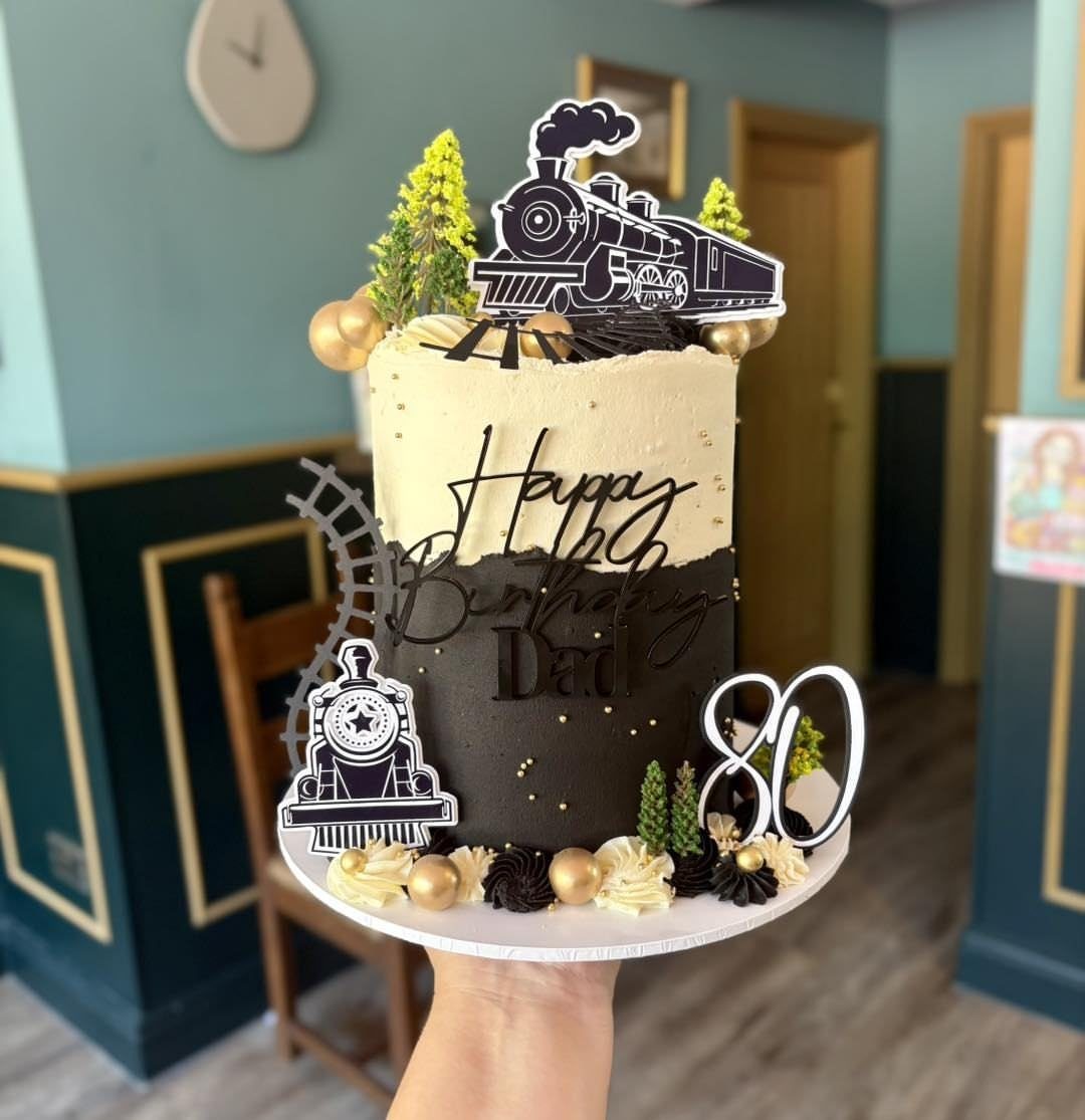 Train-themed 80th birthday cake with black and cream frosting and locomotive toppers.
