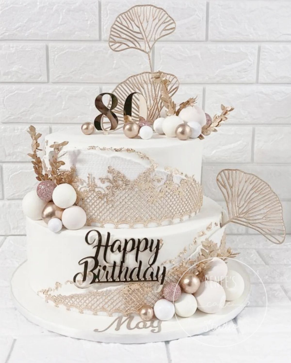 Two-tier white birthday cake with gold edible lace, gold ginkgo leaves, and 80 topper.