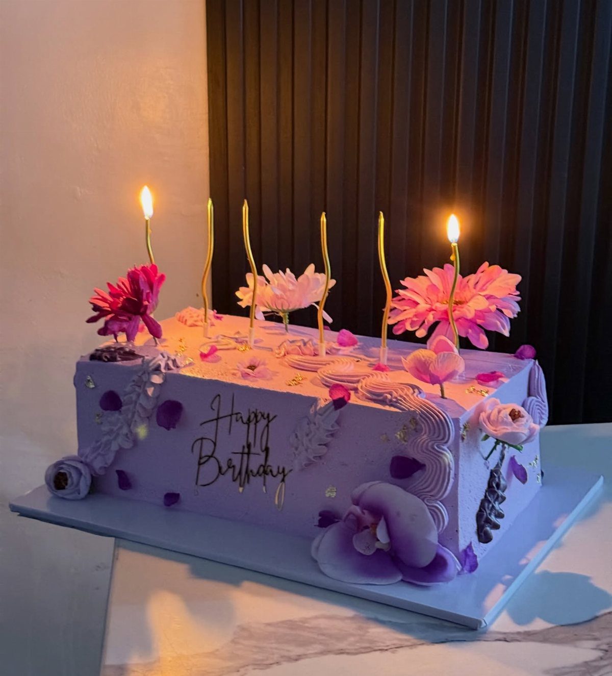 Purple birthday sheet cake decorated with pink flowers, gold candles, and gold lettering.