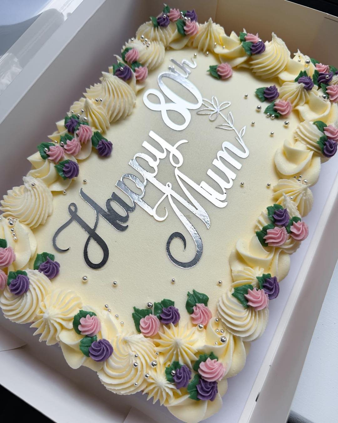 Rectangular sheet cake with yellow frosting, colorful icing flowers, and Happy 80th Mum topper.