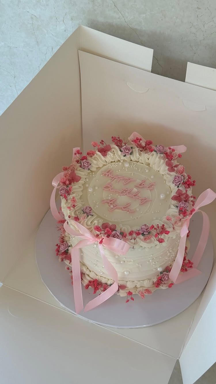 White frosted cake decorated with pink flowers, pearls, and pink ribbons in a box