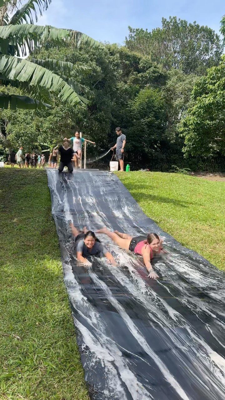 Two girls sliding down a long wet plastic slip and slide on grass.