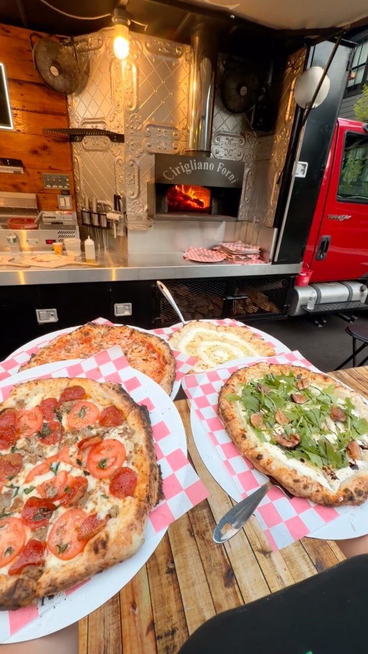 Three gourmet pizzas on a wooden table in front of a red food truck.