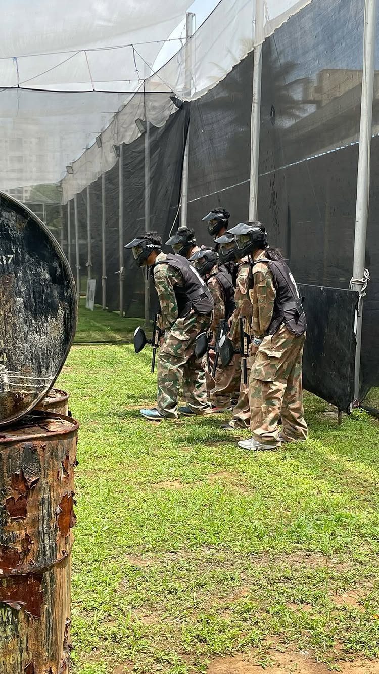 Young men in camouflage gear and helmets standing in a paintball arena.