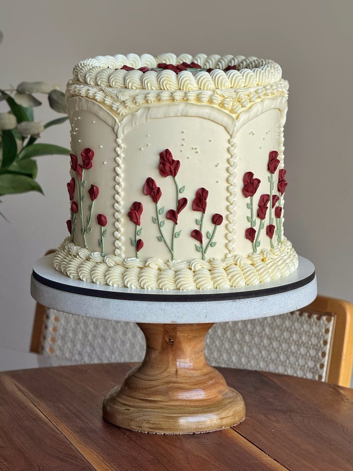 Cream-colored birthday cake decorated with red sugar flowers on a wooden stand.