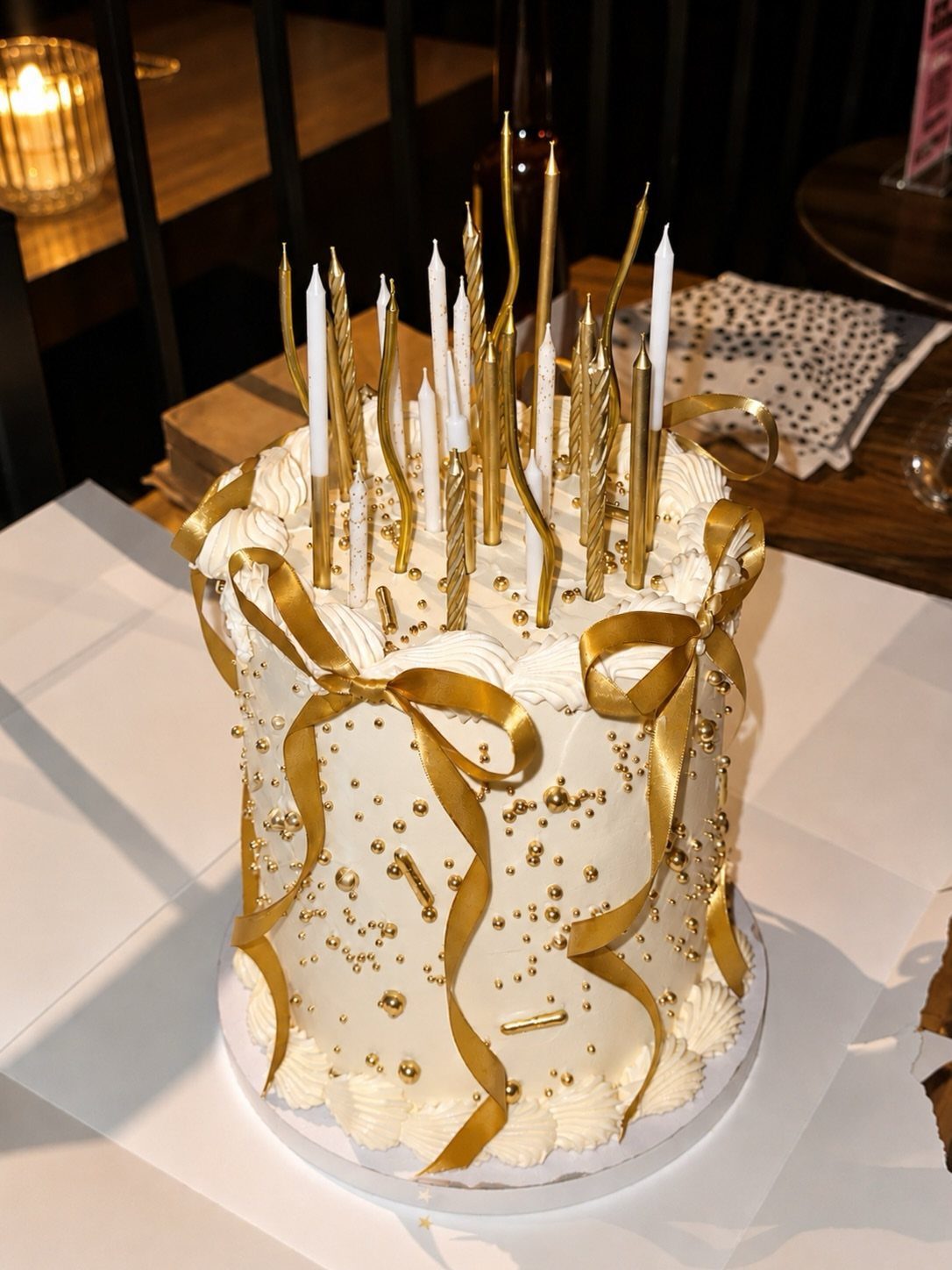 White frosted cake with gold ribbons, gold sprinkles, and numerous gold and white candles.