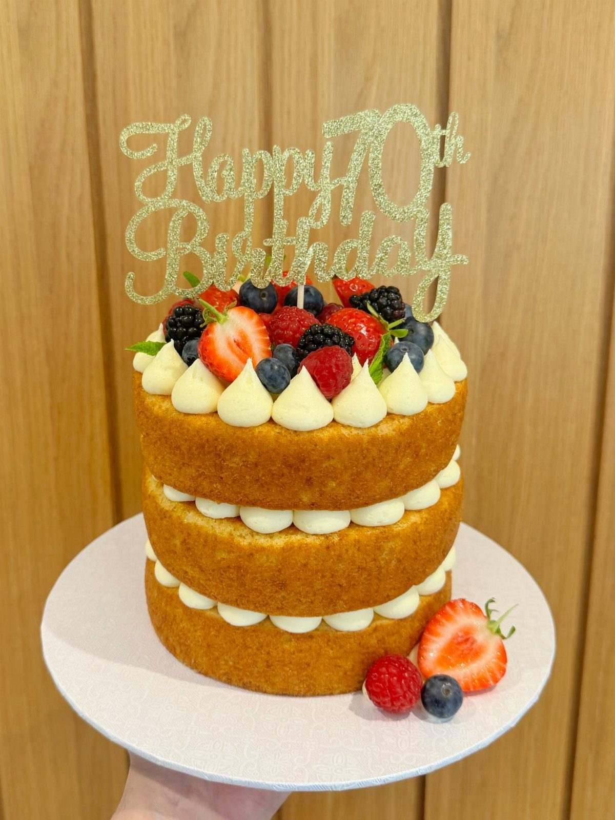 Three-layer naked cake with fresh berries and a gold Happy 70th Birthday topper.